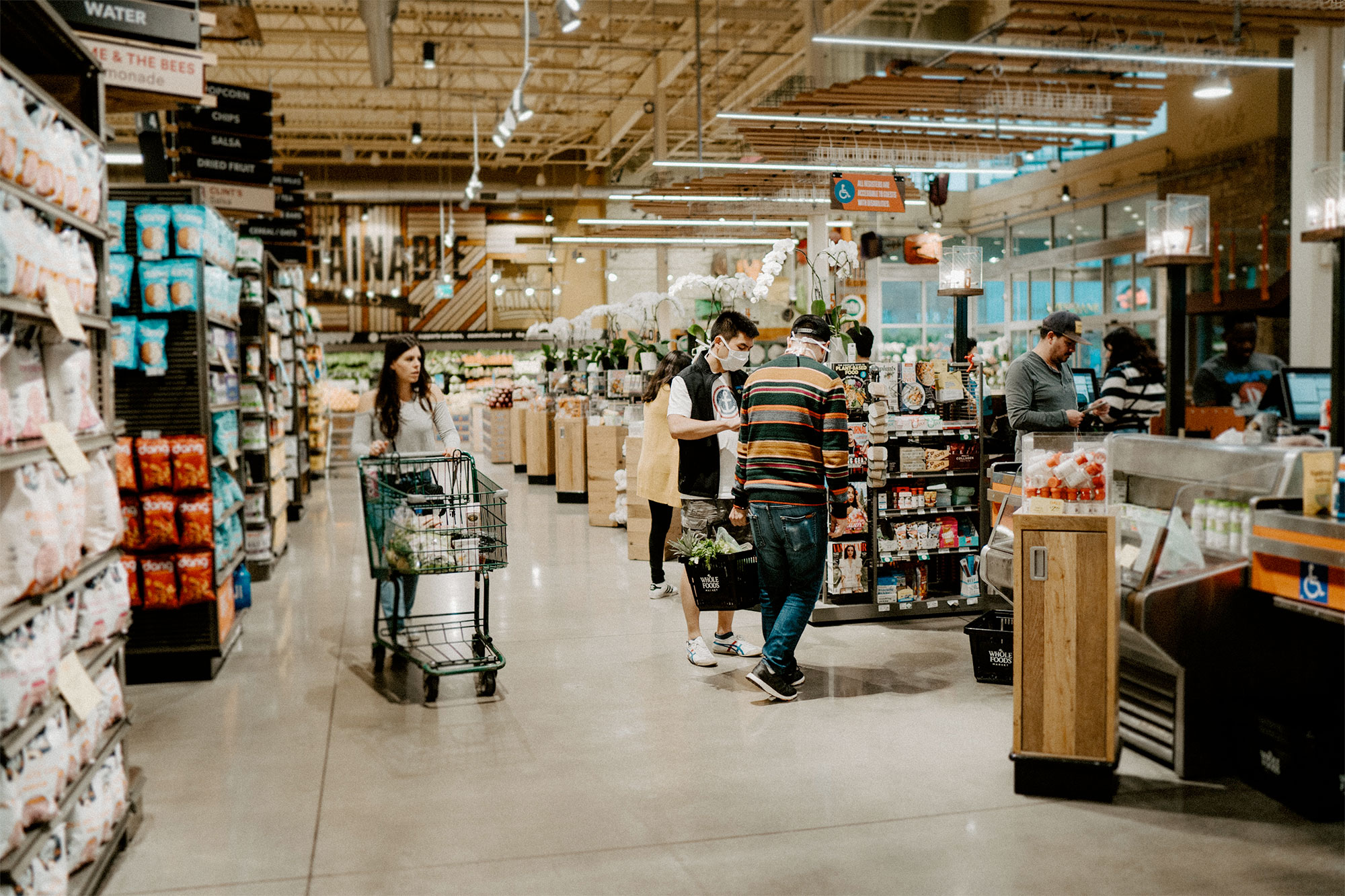 a woman walking with a shopping cart in a grocery store with people wearing medical face masks nearby